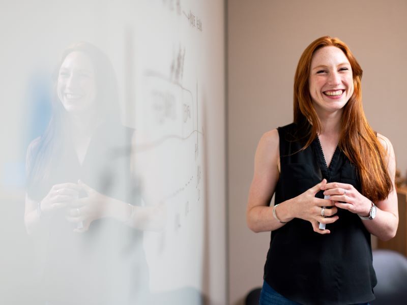 [4-3] A white female teacher stands at the front of a classroom in front of a whiteboard and holding a whiteboard pen, smiling